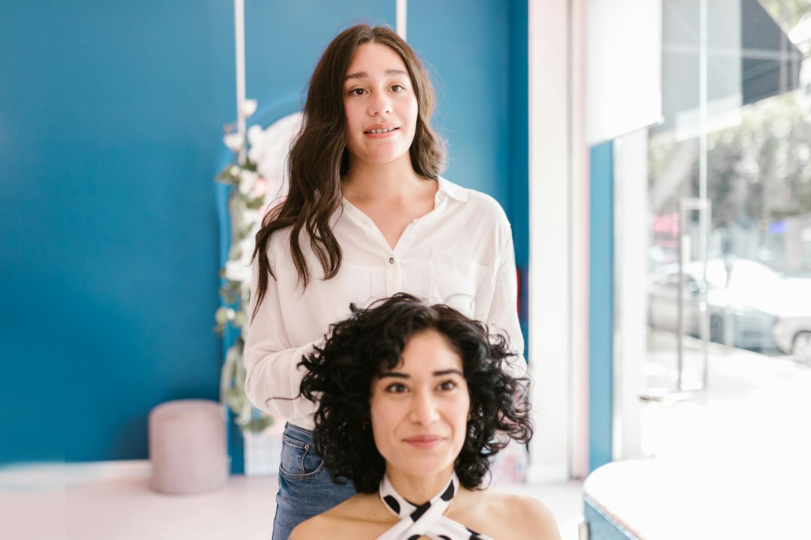 Two women in a hair salon, one styling the other's hair, both smiling brightly.